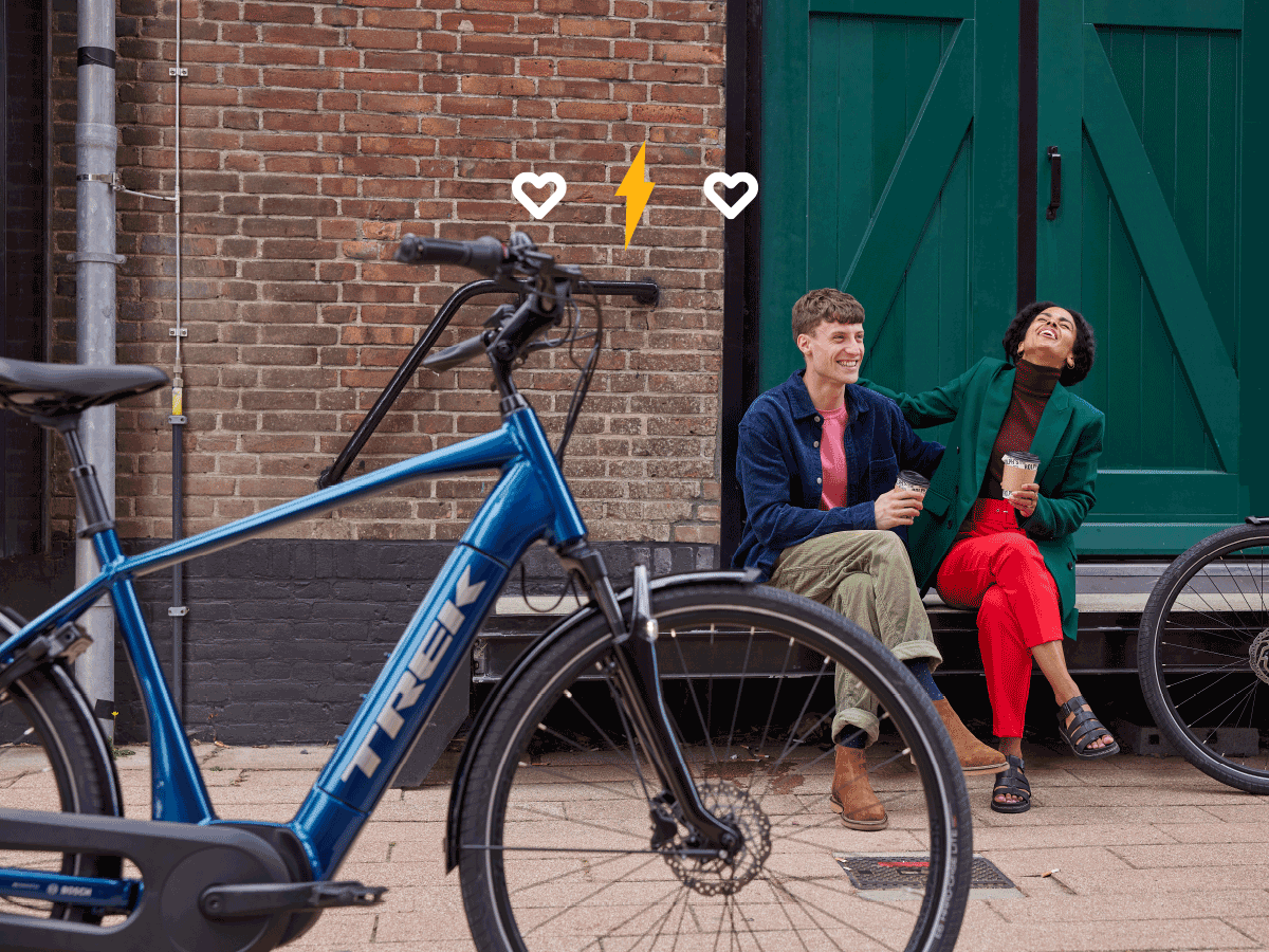 Un relámpago animado aumenta en tamaño en una ilustración de dos corazones superpuestos a una imagen de dos ciclistas sentados, riendo y tomando
    café frente a un edificio de ladrillo con una puerta verde y bicicletas Trek.