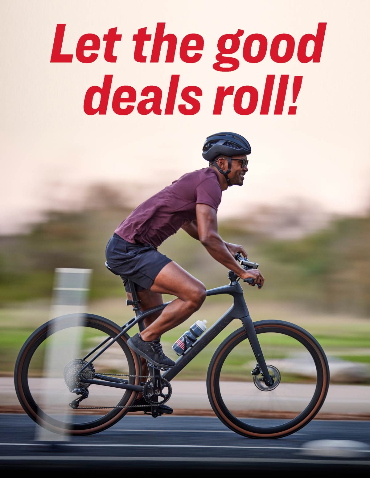 The words, 'Let the good deals roll!' overlaying image of cyclist wearing helmet and sunglasses while riding bike on paved road with trees in background
