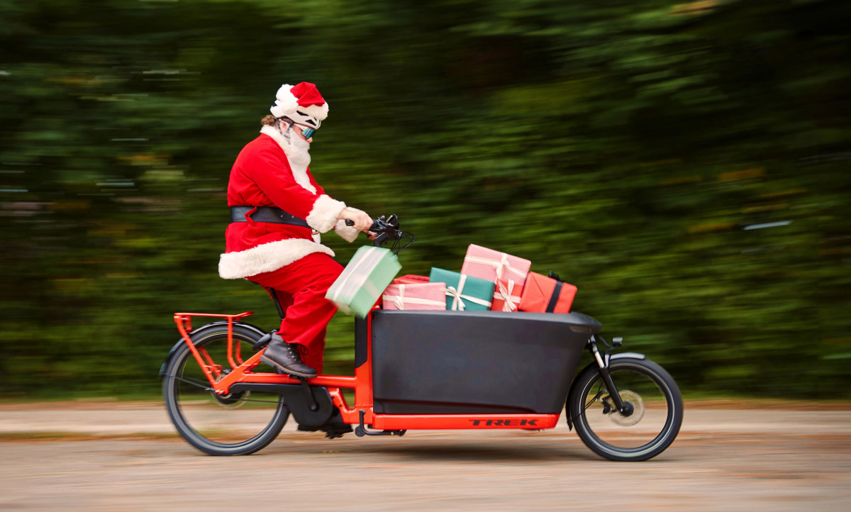 Santa riding cargo bike full of presents on paved road with trees in background