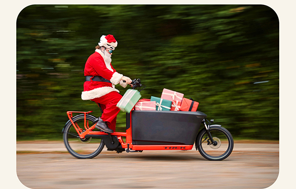 Santa riding cargo bike full of presents on paved road with trees in background