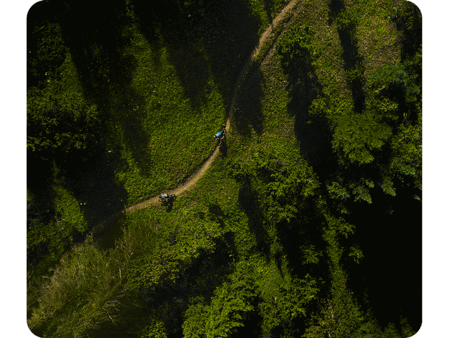 Overhead shot of two cyclists riding down a dirt trail surrounded by bright green grass and green trees