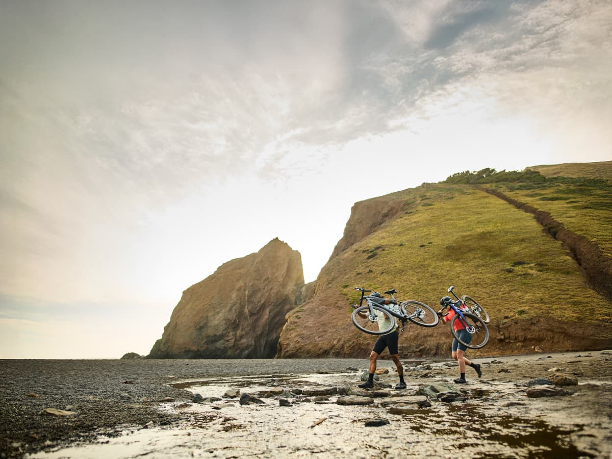 Two cyclists wearing helmets and cycling gear carrying bikes on their back, walking on rocks over a stream with a lush hill in the distance and a cloudy sunlit sky