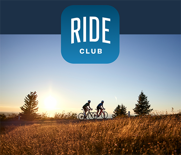 Ride Club logo overlaying photo of cyclists wearing helmets while riding bikes with tall grass in foreground and trees in background